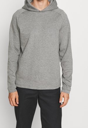 Man die een effen grijze capuchontrui met lange mouwen en een zwarte broek draagt, staand tegen een lichte achtergrond.