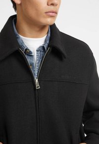 Juodas zip-up džemperis su klasikiniu apykakle ir tekstūruota medžiaga, dėvimas virš džinsinio marškinio. Ant krūtinės įspaustas logotipas "GUESS JEANS".
