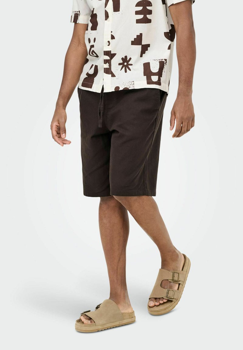 Homme portant un short noir au niveau des genoux, une chemise à manches courtes à motifs et des sandales beiges à boucles, marchant sur un fond clair uni.