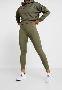 Kobieta w oliwkowych legginsach Tommy Sport i pasującym hoodie z białymi butami sportowymi, stojąca z rękami na biodrach na tle białej ściany.