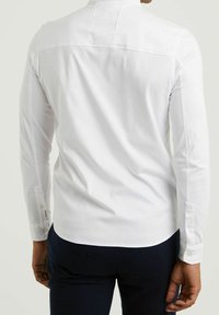 Biała koszula z długim rękawem z kołnierzykiem zapinanym na guziki, z subtelnymi detalami przeszycia i zaokrąglonym dołem, wykonana z gładkiego materiału.