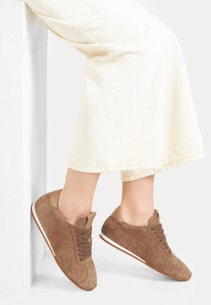 Osoba oblečená v béžových širokých kalhotách a hnědých semišových teniskách na šněrování, nohy zkřížené a opírající se o bílou zeď.