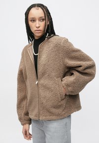Bruine shearling jas met ritssluiting, oversized pasvorm, zijzakken en beige accenten, gecombineerd met een zwarte top en grijze spijkerbroek.