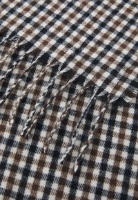 Woven plaid scarf in brown, white, and navy, featuring fringed edges and a soft texture. Checkered pattern with varying square sizes.