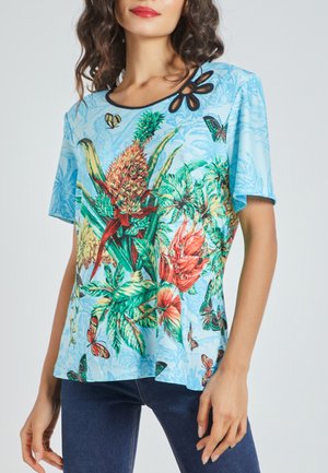 Mujer con top de manga corta azul claro con estampados coloridos de flores tropicales y mariposas y detalle decorativo negro en el escote.