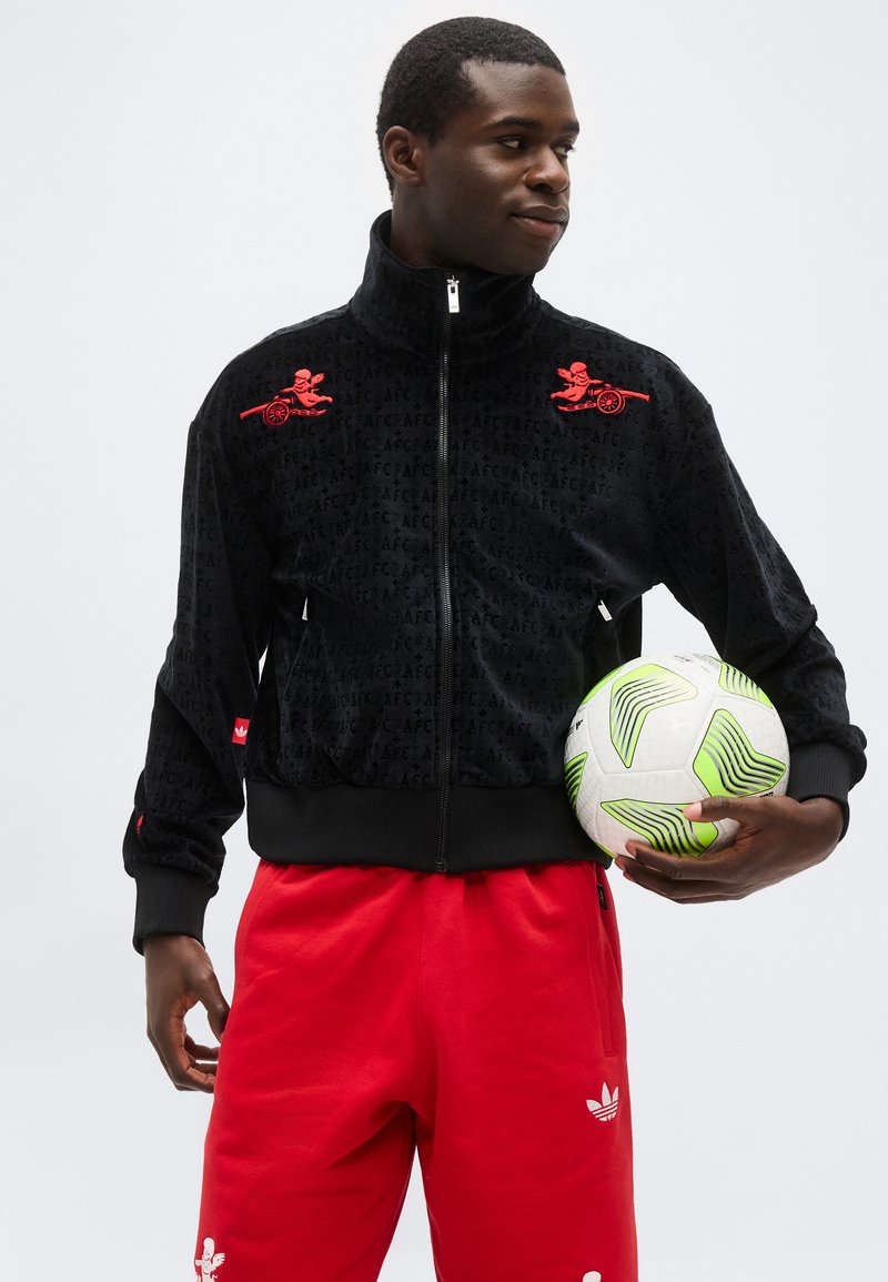 Jonge man in een zwart jack met rits met rode logo's en rode broek, die een wit-groene voetbal vasthoudt en opzij kijkt.