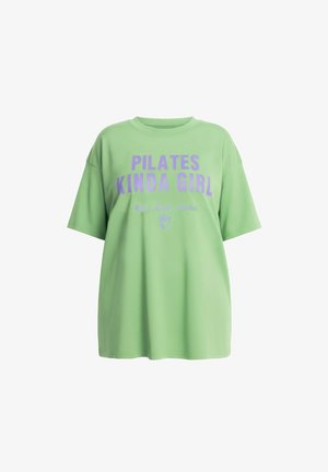 T-shirt verde chiaro oversize con scritta viola "PILATES KINDA GIRL" e una piccola illustrazione di bevanda sotto.