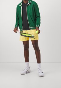 Groene jack met rits en witte accenten, zwart T-shirt, gele shorts, met een tennisracket en bal in de hand, gecombineerd met witte Nike-schoenen.