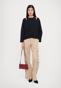 Donna che indossa un maglione nero con ritagli, pantaloni cargo beige, tacchi neri e tiene una piccola borsa rossa su uno sfondo bianco semplice.