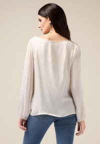 Lys beige bluse med lange ærmer, blødt stof, en rund tilbagehalsudskæring og let indsamlede skuldersømme, parret med blå jeans.