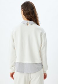Sudadera blanca con un cuello y dobladillo a rayas. Presenta un detalle de ojal en la parte posterior y una tela suave y texturizada. Mangas largas y corte relajado.