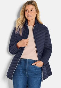 Marineblauwe gewatteerde jas met een staande kraag, ritssluiting aan de voorkant en contrasterende witte accenten. Draagt over een lichte top en denim jeans.