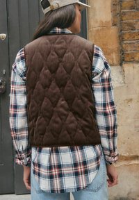 Person mit beigem Hut, braunem Steppweste, kariertem Hemd in Weiß, Marineblau und Rost sowie hellblauen Jeans, der nahe einer Backsteinmauer und einer grauen Tür steht.