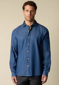 Mężczyzna w niebieskiej koszuli dżinsowej z długim rękawem, zapinanej na guziki, z kołnierzykiem, stojący na jednolitym beżowym tle.