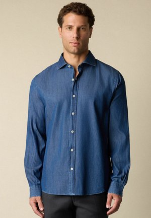 Mężczyzna w niebieskiej koszuli dżinsowej z długim rękawem, zapinanej na guziki, z kołnierzykiem, stojący na jednolitym beżowym tle.