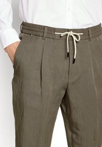 Lightweight, olive green linen trousers with a drawstring waistband, pleated front, and side pockets. Textured fabric with a relaxed fit.
