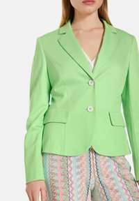 Blazer ajusté vert avec deux boutons, col cranté et poches avant. Tissu lisse avec une coupe cintrée.