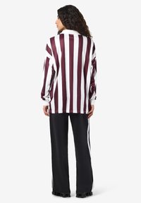 Vrouw die met haar rug naar de camera staat, gekleed in een langmouwen shirt met verticale bordeauxrode en witte strepen en zwarte wijde broek met witte zijstreep.
