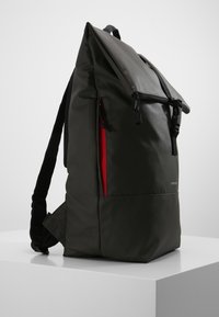 Dark green backpack with a fold-over top, textured fabric, black straps, and a red zippered pocket on the side. Simple logo detail.