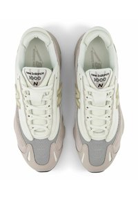 New Balance 1000 -kengät ovat vaalean kerman väriset, joissa on harmaa verkko ja vaaleanpunaisia yksityiskohtia, pyöreät nauhat ja pehmustettu pohja.