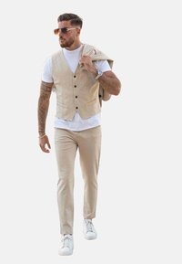 Gilet en lin beige à cinq boutons sur un T-shirt blanc, associé à un pantalon beige clair et des baskets blanches ; lunettes de soleil, montre au poignet.