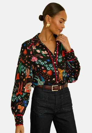 Frau mit Dutt, die eine schwarze Bluse mit Blumenmuster und rotem Rand, hochtaillierte dunkle Jeans, goldene Ohrringe und einen braunen Gürtel trägt, posiert vor weißem Hintergrund.