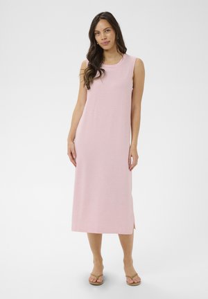 Kobieta o długich ciemnych włosach, ubrana w różową, bezrękawnikową sukienkę do połowy łydki oraz beżowe sandały, stojąca na białym tle.