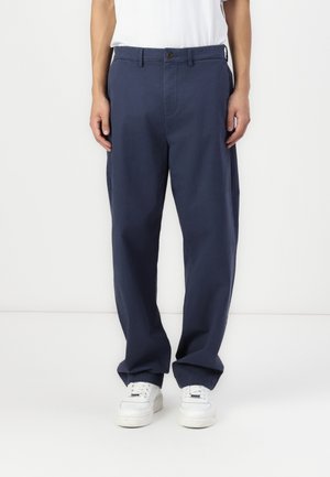 Mężczyzna ubrany w granatowe, luźne spodnie i białe tenisówki, stojący na tle jednolitego białego tła.