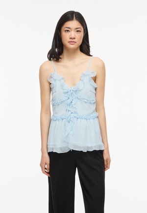 Žena má na sebe svetlomodrú volánikovú blúzku bez rukávov s tenkými ramienkami a čierne nohavice, stojí pred bielym pozadím.