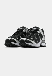 Ténis preto e branco com parte superior em malha e sola de borracha, apresentando um design curvo, acabamentos texturizados e um sistema de atilhos integrado.