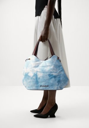 Personne tenant un grand sac à main tie-dye bleu et blanc avec des poignées marron, portant une jupe blanche et des escarpins noirs à bout pointu.