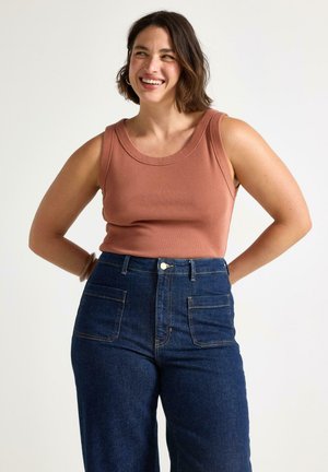 Smilende kvinde med kort brunt hår iført en ærmeløs rustfarvet top og mørkeblå jeans med høj talje og forlommer.