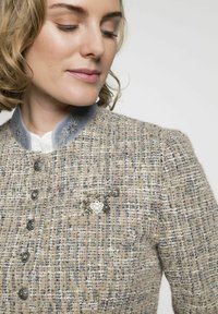 Tweedjacke in Beige-Tönen, mit silbernen Knöpfen und einem blauen Kragen. Verfügt über eine dekorative Brosche mit einem Herzen und Tiermotiven. Strukturierter Stoff.
