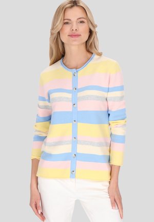 Vrouw draagt een pastel gestreepte gebreide cardigan met knopen, met gele, roze, blauwe, witte en grijze strepen en witte broek, staand tegen een effen achtergrond.
