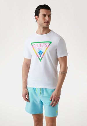 White cotton t-shirt featuring a multicolored triangle logo with palm tree and "GUESS BEACH" text; paired with light blue swim shorts.