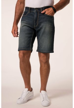 Mand iført mørkeblå denimshorts, hvide sneakers og en hvid skjorte med den ene hånd i lommen og den anden afslappet langs siden.