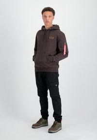 Sudadera con capucha de tejido marrón oscuro, con bolsillo frontal, cordones y un bolsillo con cremallera lateral que presenta una etiqueta roja; combinada con pantalones cargo negros.
