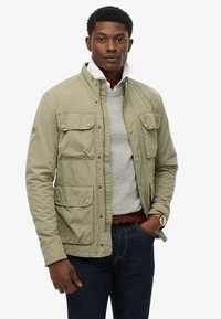 Olijfgroene jas met een rits en drukknopen, vier voorzakken en een hoge kraag over een grijze trui. Getextureerde stof, casual pasvorm.