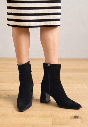 Botas de tornozelo em camurça preta com bico em ponta e salto quadrado robusto. Apresenta um zíper exterior ao longo do lado.