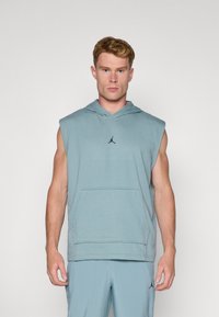 Ærmeløs hoodie i lyseblåt stof. Har en kengurulomme og et sort logo på brystet. Simpelt, udekoreret design.
