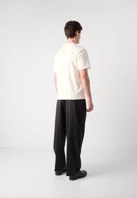 Light beige short-sleeve t-shirt with a crew neck, paired with loose black trousers and black shoes, viewed from the back.