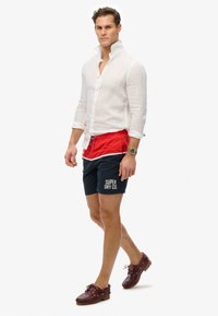 Camisa blanca de lino de botones, bañador rojo y azul marino con el logo "SUPER DRY CO.", zapatos de barco marrones, en pose de pie contra un fondo sencillo.