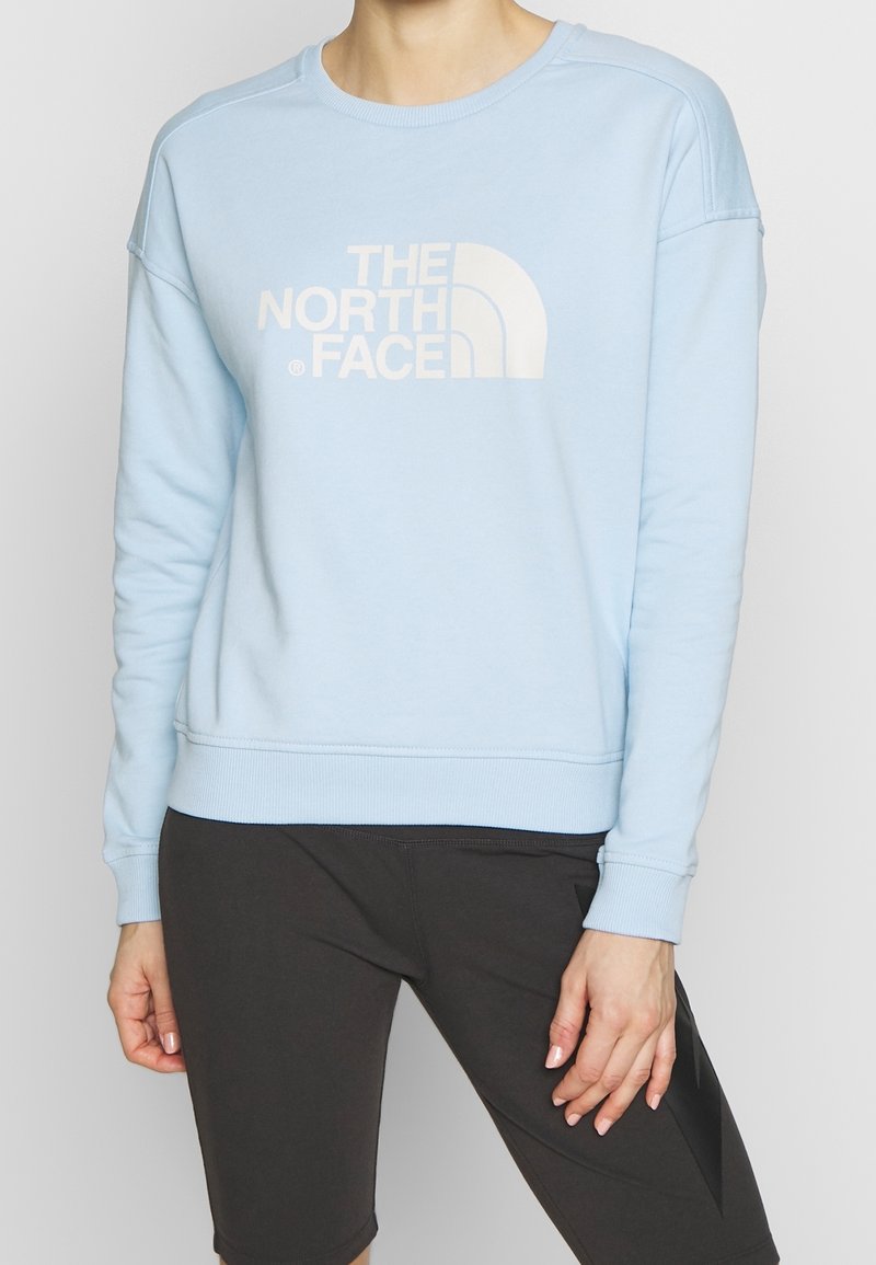 Lyseblå sweatshirt med en stor hvid logo med et delvist buet design. Fremstillet af blødt stof med rund halsudskæring og ribbede manchetter.