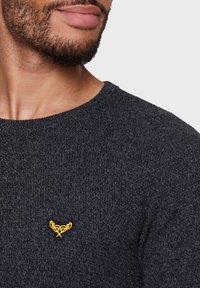 Pull en maille gris foncé avec un col rond et un petit logo décoratif doré sur la poitrine. Texture douce avec une apparence confortable.