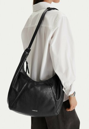 Mujer con camisa blanca lleva un gran bolso de hombro de cuero negro con el logo visible de la marca "Lasocki".