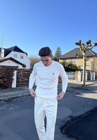 Tânăr îmbrăcat într-un pulover alb cu model tricotat în relief și pantaloni crem, stând pe o stradă rezidențială liniștită, cu case și un copac tuns sub un cer senin, albastru.