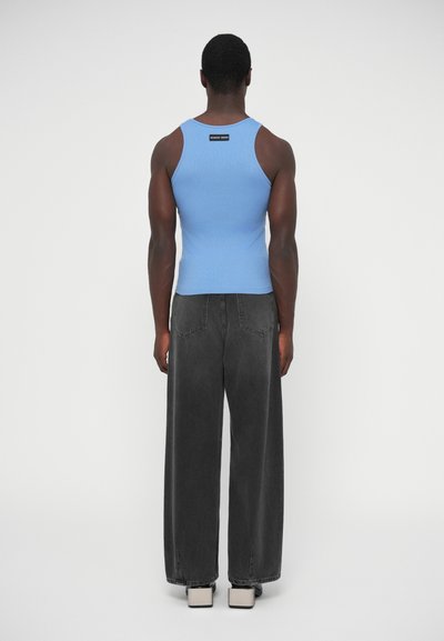 Modrý rebrovaný tielko s logom na zadnej strane, v kombinácii s voľnými sivými džínsami. Oblečenie doplňujú neutrálnych farieb topánky.