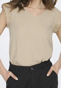 Blouse beige avec un col en V et des manches en dentelle ajourée festonnée, associée à un pantalon noir taille haute avec une fermeture à boutons.