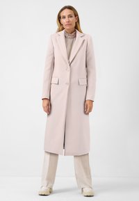 Ljusrosa skräddarsydd kappa med lång silhuett, tvåknapps-stängning och sidfickor. Bärs över en plisserad topp och parat med ljusa byxor.