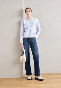 Lyse lilla blomstret genser, tettsittende i midjen, sammen med mørkeblå rette jeans. Holder en kremfarget håndveske og har på seg kremfargede platåsko.